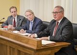 Mike Freeman sits at the testifiers' table and speaks to someone out of view. Representative Dehn and Secretary of State Steve Simon sit next to him and watch as he speaks.